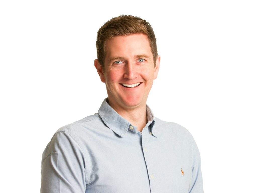 A man with short brown hair is smiling at the camera. He is wearing a light blue, button-down shirt and stands in front of a plain white background.