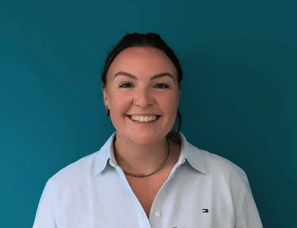 A person with dark hair pulled back, wearing a light blue collared shirt and a chain necklace, smiles at the camera against a solid teal background.