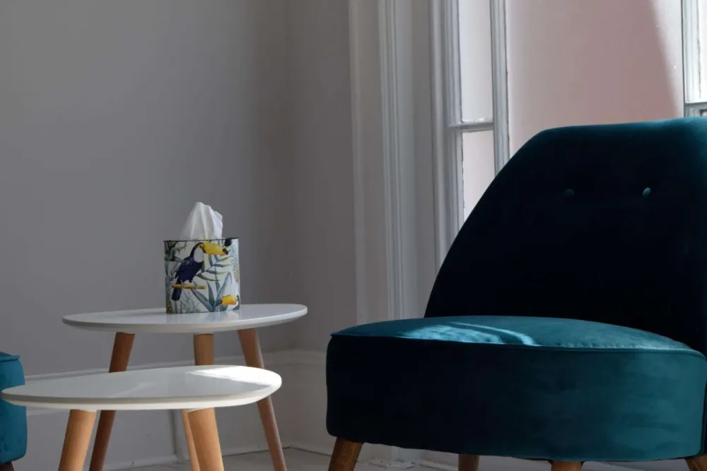 A cozy corner featuring a dark teal velvet chair next to a round white side table. The table has a tissue box with a colorful floral pattern on top. Soft natural light filters in from a nearby window, creating a serene ambiance.