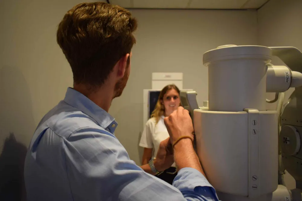 A man with brown hair, wearing a blue shirt, adjusts medical imaging equipment. A woman with blonde hair, wearing a white shirt, stands in front of the machine, preparing for a scan in a clinical setting.