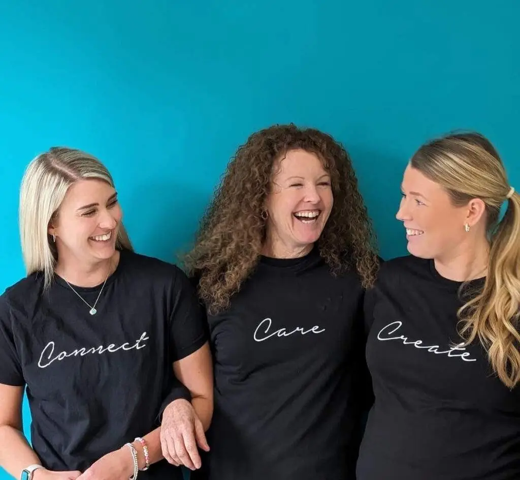 Three women Charlotte Page, Sharon Johnstone, Sara Egerfors from the Life Balance Chiropractic Team stand closely together, smiling and laughing against a blue background. Each woman wears a black t-shirt with white text: the shirts read "Connect," "Care," and "Create" from left to right. They appear to enjoy each other's company.