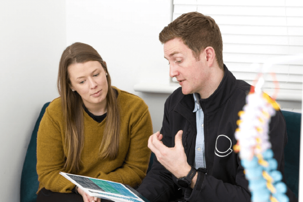 A man and a woman are sitting together, engaged in a discussion. The man, who appears to be a chiropractor, is gesturing and holding a pamphlet, explaining something to the woman. She is listening attentively. There is part of a colorful anatomical model in the foreground, likely used at their chiropractic clinic.