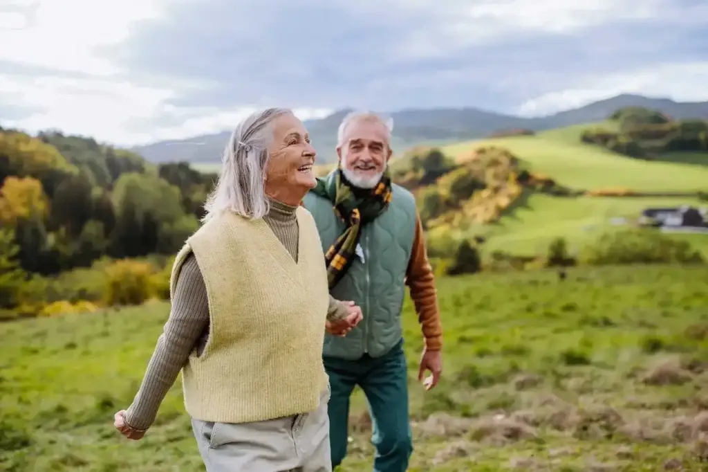 Christchurch Chiropractor 2 An elderly couple enjoys a sunny day in the countryside. The woman, wearing a yellow vest over a brown turtleneck, walks ahead with a smile. The man, dressed in a green jacket and scarf, follows behind. Green fields and hills stretch out in the background.